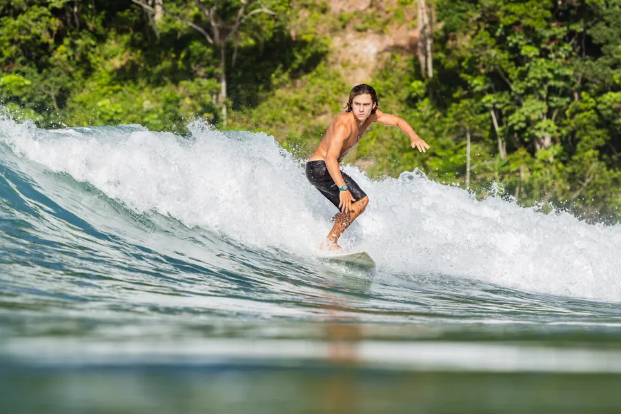 Andre desce a onda com o braço estendido, mata verde ao fundo